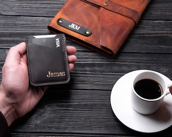 May include: A brown leather card holder with the name "James" embossed in gold lettering. The card holder is sitting on a dark wooden surface.