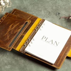 May include: A brown leather planner with a yellow accent and a zippered pocket. The planner is open to a page with the word "PLAN" printed on it.