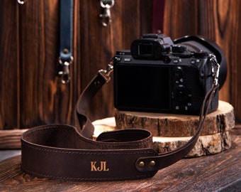 May include: Brown leather camera strap with the initials "KJL" embossed in gold. The strap is attached to a black camera with a lens.