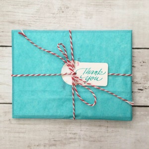 Pode incluir: Uma embalagem retangular embrulhada em papel de seda turquesa e amarrada com barbante vermelho e branco. Uma pequena etiqueta com as palavras "Thank You" est&aacute; presa ao barbante. A embalagem est&aacute; sobre uma superf&iacute;cie de madeira clara.