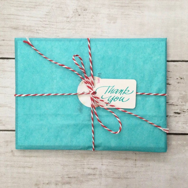 Pode incluir: Uma embalagem retangular embrulhada em papel de seda turquesa e amarrada com barbante vermelho e branco. Uma pequena etiqueta com as palavras "Thank You" est&aacute; presa ao barbante. A embalagem est&aacute; sobre uma superf&iacute;cie de madeira clara.