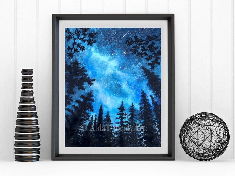 May include: A framed watercolor painting of a night sky with a bright blue and white galaxy, silhouetted trees, and a shooting star. The artwork is signed "Aida Tubic Art". A black decorative vase and a wire sphere are on the white surface.
