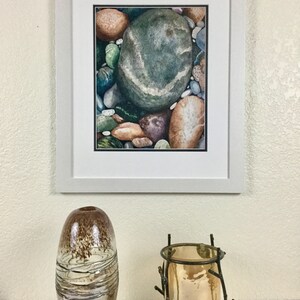 May include: A framed watercolor painting of colorful river rocks, including a large green stone with a white stripe. Below, a decorative brown and silver glass vase and a metal candle holder with a gold-colored candle.