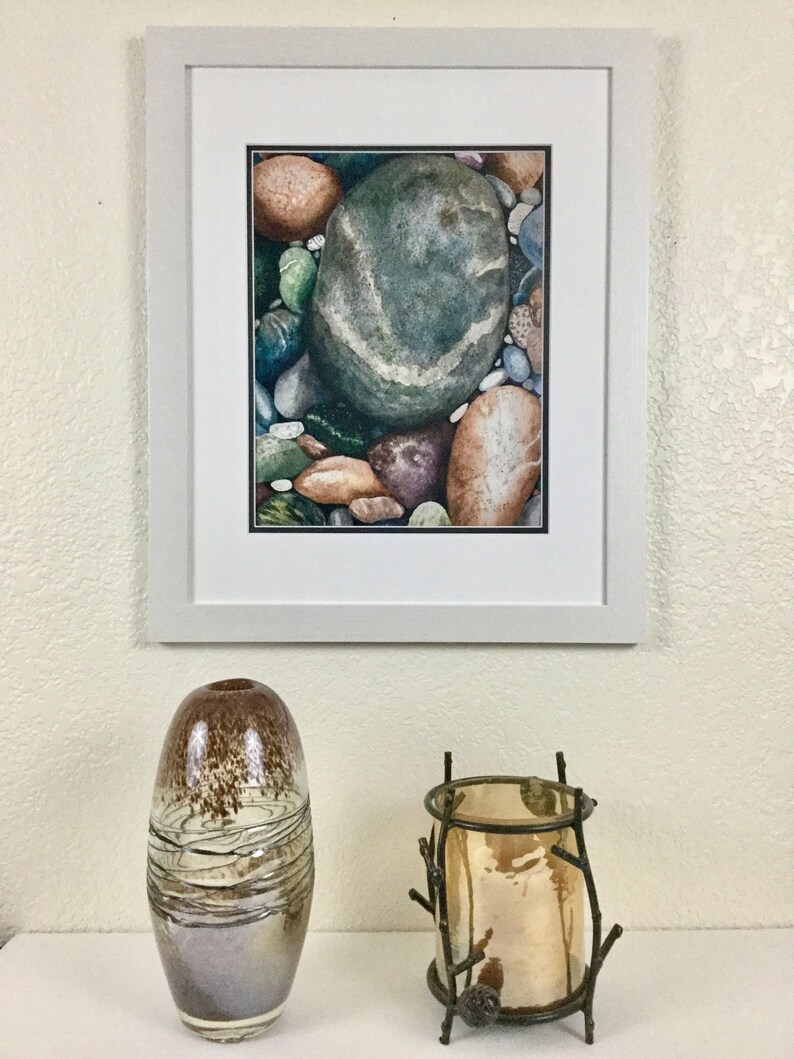 May include: A framed watercolor painting of colorful river rocks, including a large green stone with a white stripe. Below, a decorative brown and silver glass vase and a metal candle holder with a gold-colored candle.