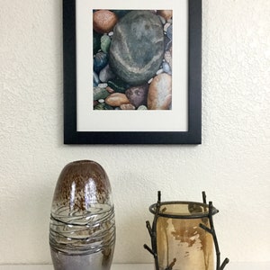 May include: A framed watercolor print of river rocks hangs above a decorative display. The display includes a brown and silver glass vase and a bronze candle holder with a glass insert.