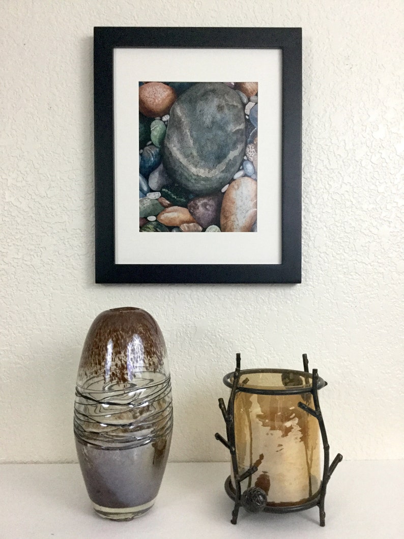 May include: A framed watercolor print of river rocks hangs above a decorative display. The display includes a brown and silver glass vase and a bronze candle holder with a glass insert.