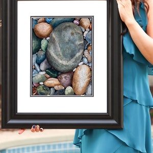 May include: A framed art print featuring a detailed watercolor painting of various smooth river rocks. The rocks are in shades of brown, gray, blue, and green, with a large, oval gray stone dominating the center. The frame is black.