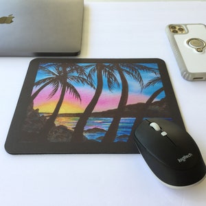 Pode incluir: Um mousepad retangular preto com uma cena colorida de p&ocirc;r do sol com palmeiras. Um mouse sem fio preto e um laptop est&atilde;o ao lado do mousepad. Um smartphone com um suporte de anel tamb&eacute;m est&aacute; na imagem.