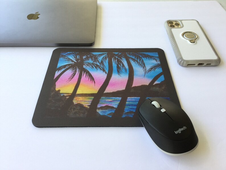 Pode incluir: Um mousepad retangular preto com uma cena colorida de p&ocirc;r do sol com palmeiras. Um mouse sem fio preto e um laptop est&atilde;o ao lado do mousepad. Um smartphone com um suporte de anel tamb&eacute;m est&aacute; na imagem.
