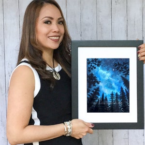 May include: A framed watercolor painting of a night sky with a blue and white galaxy, silhouetted trees, and a few stars. The artwork is in a black frame with a white mat.