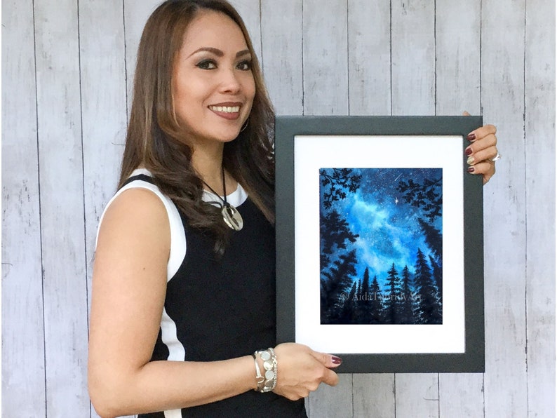 May include: A framed watercolor painting of a night sky with a blue and white galaxy, silhouetted trees, and a few stars. The artwork is in a black frame with a white mat.