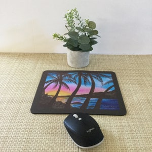 Pode incluir: Tapete de rato retangular preto com uma cena de p&ocirc;r do sol colorida com palmeiras, oceano e c&eacute;u em tons de azul, rosa e amarelo. Um rato de computador preto est&aacute; posicionado abaixo. Uma pequena planta em vaso est&aacute; no fundo.