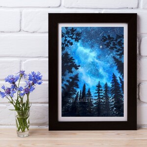 May include: A framed watercolor painting depicts a night sky with a vibrant blue and white galaxy, silhouetted trees, and a few stars. The artwork is in a black frame. A small glass vase with blue flowers sits to the left.