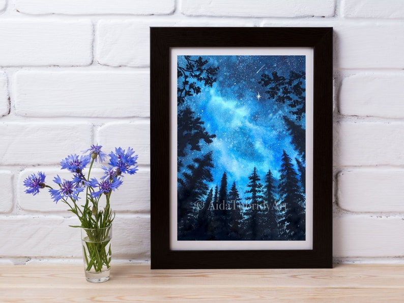 May include: A framed watercolor painting depicts a night sky with a vibrant blue and white galaxy, silhouetted trees, and a few stars. The artwork is in a black frame. A small glass vase with blue flowers sits to the left.