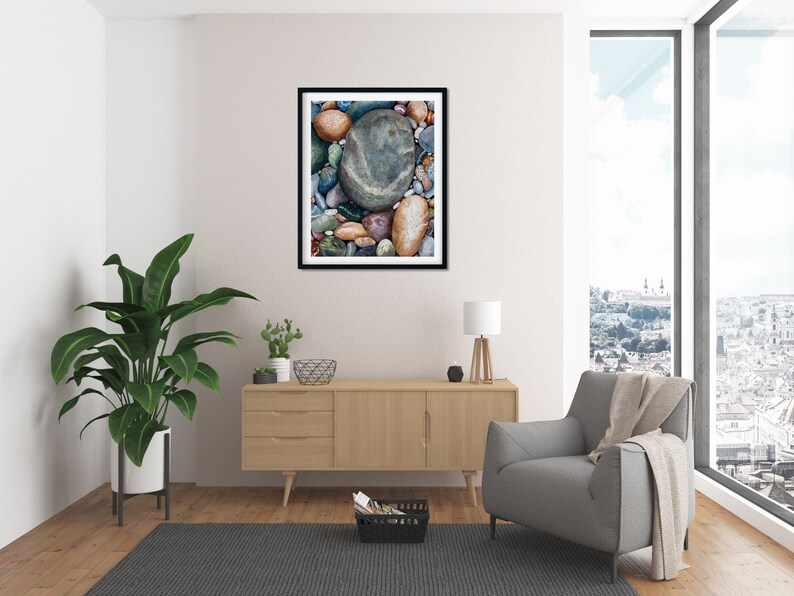 May include: A framed print featuring a collection of smooth, colorful river rocks. The rocks vary in size and color, with shades of brown, blue, green, and gray. The artwork is displayed in a modern living room setting with a large window and contemporary furniture.