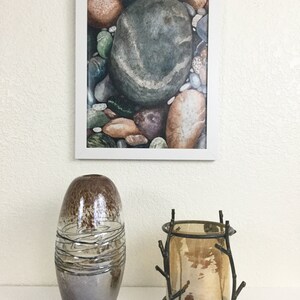 May include: A framed watercolor print of colorful river rocks hangs above a decorative display. Below, a brown and silver glass vase with swirling lines and a bronze candle holder with a glass insert and twig-like design.