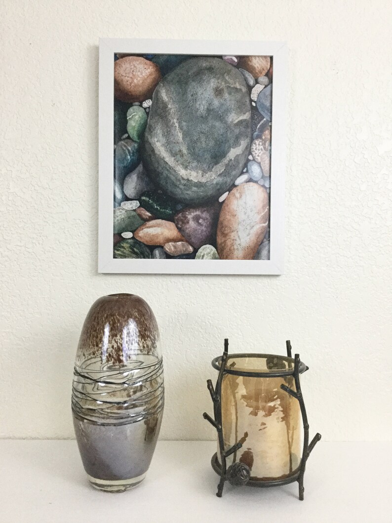 May include: A framed watercolor print of colorful river rocks hangs above a decorative display. Below, a brown and silver glass vase with swirling lines and a bronze candle holder with a glass insert and twig-like design.
