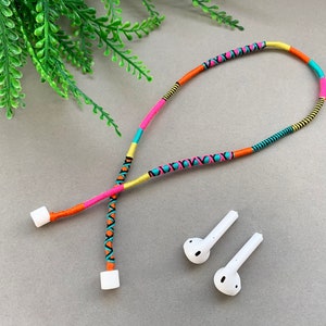 Puede incluir: Un cordón trenzado de colores con extremos de plástico blanco, diseñado para evitar que los auriculares se enreden. También se muestran dos auriculares inalámbricos blancos.