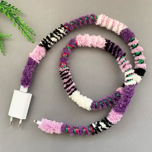 May include: A white phone charger cord wrapped in a colorful yarn with purple, pink, white, black, and green stripes.