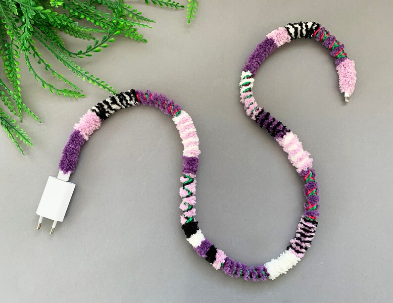 May include: A white phone charger cord with a purple, pink, white, black, and green yarn wrap.