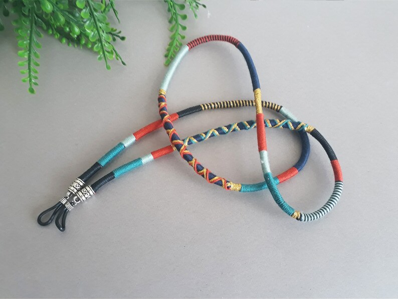May include: A colorful braided eyeglass chain with black cord ends and silver metal clasps. The chain is made of blue, red, green, and gold threads.