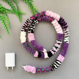 May include: A colorful phone charging cable wrapped in yarn with purple, pink, white, black, and green stripes. The cable is coiled and ready to be used.