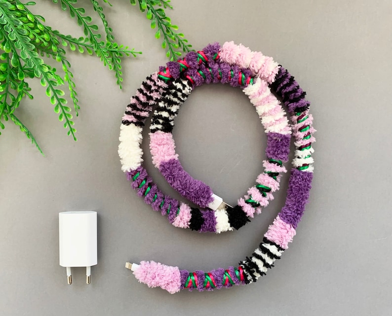 May include: A colorful phone charging cable wrapped in yarn with purple, pink, white, black, and green stripes. The cable is coiled and ready to be used.