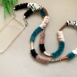 May include: A colorful phone strap made of fuzzy yarn in teal, black, white, and brown. The strap is attached to a clear phone case.