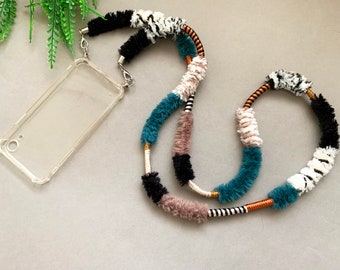 May include: A colorful phone strap made of fuzzy yarn in teal, black, white, and brown. The strap is attached to a clear phone case.