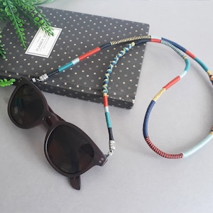 May include: A pair of black sunglasses with a colorful braided cord. The cord is made of blue, red, yellow, and white thread. The sunglasses are lying on a gray surface.