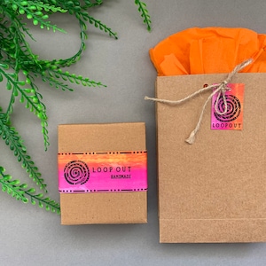 May include: Two brown cardboard boxes with a pink and orange graphic design. The smaller box has the text "LOOP OUT HANDMADE" on it. The larger box has a tag with the text "LOOP OUT" on it.
