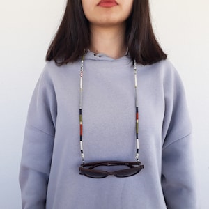 May include: A person wearing a grey hoodie with a black, white, red, and green beaded glasses chain. The chain is holding a pair of brown sunglasses.