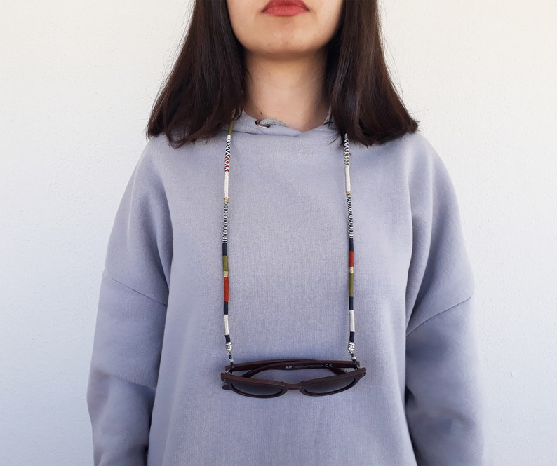 May include: A person wearing a grey hoodie with a black, white, red, and green beaded glasses chain. The chain is holding a pair of brown sunglasses.