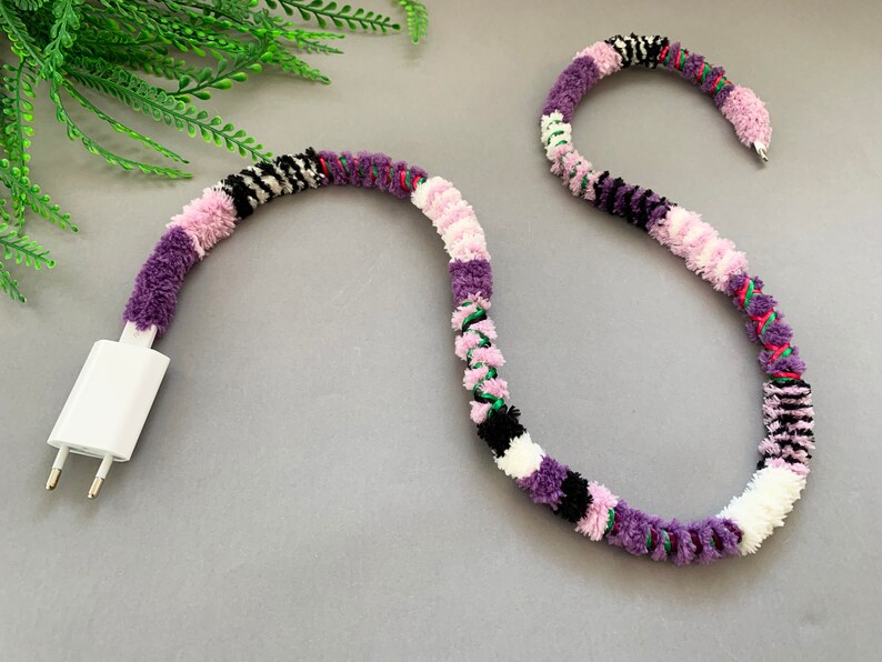 May include: A white phone charger cord wrapped in a colorful yarn with purple, pink, white, black, and green stripes.