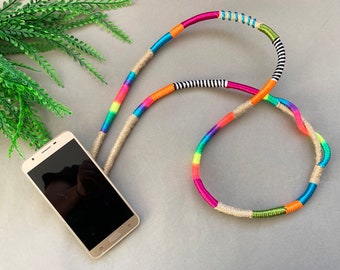 May include: A colorful phone strap made of woven yarn with a rainbow pattern. The strap is made of a natural fiber and has a brown and white color scheme. The strap is attached to a black smartphone.