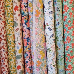 Könnte beinhalten: Gerollte Stoffballen mit verschiedenen floralen und botanischen Mustern. Die Stoffe zeigen eine Reihe von Farben, darunter Rosa, Gelb, Blau und Rot, mit Designs von Blumen, Blättern und Beeren. Die Ballen sind nebeneinander angeordnet.