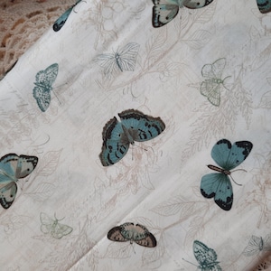 Könnte beinhalten: Cremefarbener Stoff mit einem sich wiederholenden Muster aus türkis- und braunfarbenen Schmetterlingen und floralen Elementen. Die Schmetterlinge haben detaillierte Flügelmuster, und der Stoff hat eine Vintage-Ästhetik. Geeignet für Bastelprojekte.