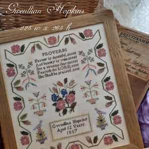 May include: A framed cross-stitch sampler with a white background and colourful floral designs. The sampler includes a verse from Proverbs and the name "Gwenllian Hopkins" with the age "12 years" and the year "1857".