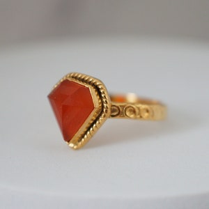 Puede incluir: Un anillo de oro con una gran piedra roja facetada. El anillo tiene una banda decorativa.