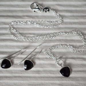 Puede incluir: Un conjunto de collar y pendientes de plata con colgantes de piedras preciosas negras. El collar tiene un cierre de plata y una cadena delicada. Los pendientes son pendientes colgantes con un gancho de plata.