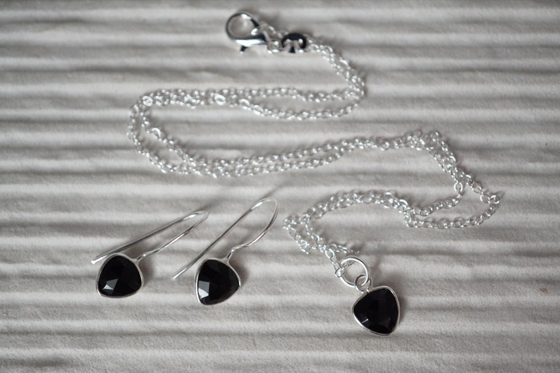 Puede incluir: Un conjunto de collar y pendientes de plata con colgantes de piedras preciosas negras. El collar tiene un cierre de plata y una cadena delicada. Los pendientes son pendientes colgantes con un gancho de plata.