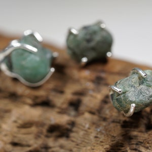 Puede incluir: Tres anillos de plata con engastes de piedras preciosas verdes. Los anillos están sobre un trozo de madera desgastada.