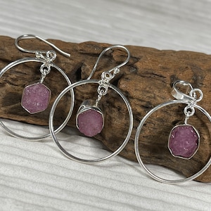 May include: A set of three silver hoop earrings with pink gemstone pendants. Each earring features a circular hoop with a dangling pink gemstone.