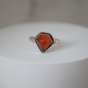Puede incluir: Un anillo de plata con una piedra preciosa naranja facetada engastada en un engaste de plata. El anillo tiene una banda torcida.