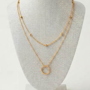 Op de afbeelding: Een goudkleurige, gelaagde ketting weergegeven op een mannequin. De bovenste laag heeft kleine kralen, terwijl de onderste laag een cirkelvormige hanger heeft. De ketting is delicaat en lijkt van metaal te zijn.