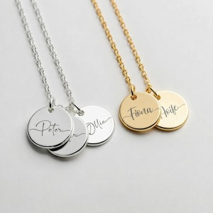 Puede incluir: Dos collares personalizados con colgantes redondos. Uno es plateado con los nombres "Peter" y "Mia" grabados. El otro es dorado con los nombres "Fiona" y "Aoife" grabados. Ambos collares tienen delicadas cadenas.
