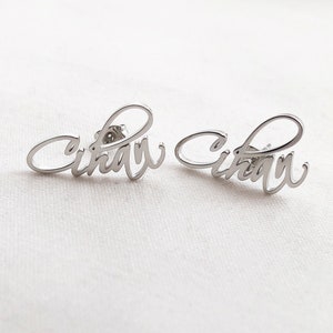Puede incluir: Un par de pendientes de plata con el nombre "Cihari" escrito en cursiva.