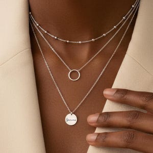 May include: A layered silver necklace set. The top layer features small beads, the middle a simple circle pendant, and the bottom a round pendant with the word "Gemini" in cursive. The necklaces are worn over a beige blazer.