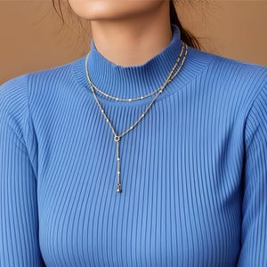 Puede incluir: Una mujer lleva un suéter azul de cuello alto de canalé con un collar de capas de oro con un pequeño colgante.