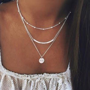 May include: A silver layered necklace set. The top layer has small beads, the middle layer a curved bar with text, and the bottom layer a round pendant with the name "Amelia". The necklaces are worn with a white top.